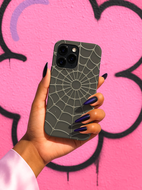 A hand holding a phone with a spider web pattern on the back cover, against a pink graffiti background.